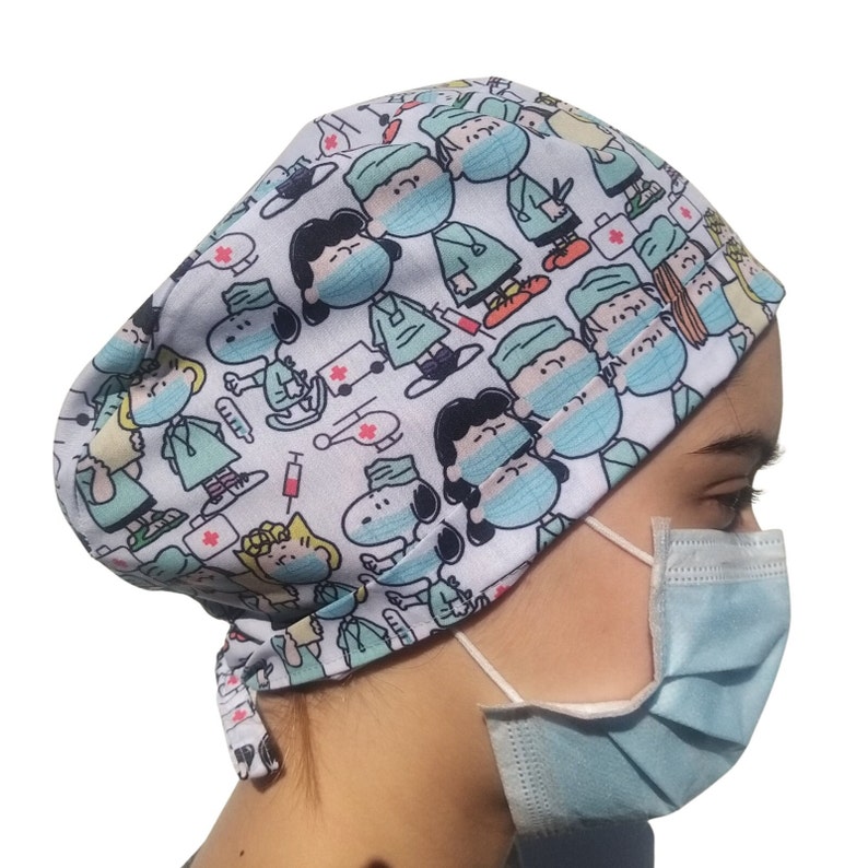 May include: A white scrub hat with a colorful print of cartoon characters wearing surgical masks and scrubs. The characters include Snoopy and Charlie Brown. The hat has a tie at the back.