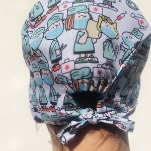May include: A white scrub hat with a blue and green pattern of cartoon characters wearing surgical masks and scrubs. The hat has a tie at the back.