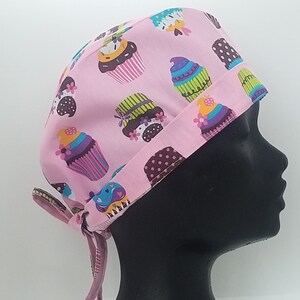 May include: Pink surgical scrub hat with a cupcake pattern. The hat has a tie on the back and a brown trim.