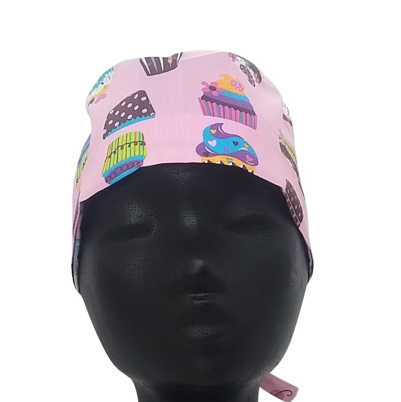 May include: Pink surgical scrub hat with a colorful cupcake pattern. The hat has a tie at the back.