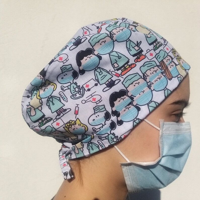 May include: A white scrub cap with a blue and white pattern of Peanuts characters wearing surgical masks and holding medical supplies. The cap has a tie at the back.