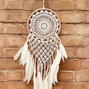 May include: A white dream catcher with a crocheted circular design and white feathers hanging from the bottom. The dream catcher is hanging on a brick wall.