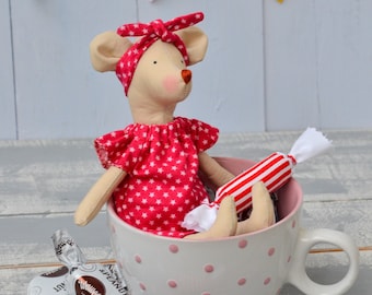 Könnte beinhalten: Eine beige Stoffmauspuppe in einem roten Kleid mit weißen Sternen und passendem Stirnband, die in einer weißen Keramiktasse mit rosa Punkten sitzt. Die Puppe hält ein rot-weiß gestreiftes Stoffbonbon, weitere Bonbons liegen daneben.