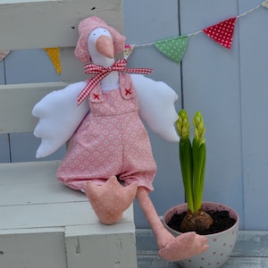 May include: A handmade goose doll in a pink floral print outfit, with overalls and a bonnet. The goose has white wings and a red and white checked bow. A potted plant with green stalks and buds is next to it. Decorative bunting hangs in the background.