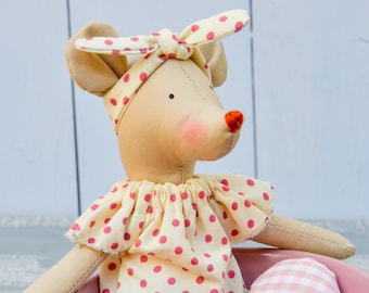 Könnte beinhalten: Eine handgefertigte beige Mäusepuppe mit roter Nase, rosa Wangen und einem gelben Stirnband und Kleid, beide mit rosa Tupfen verziert. Die Puppe sitzt in einer rosa Schüssel mit einem rosa-weiß karierten Akzent.