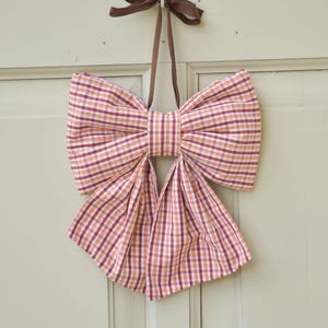 May include: A large decorative bow made of fabric with a red, orange, and white gingham checked pattern. The bow features two wide top loops and two long, flowing bottom tails, and it is suspended by a thin brown ribbon loop at the top.
