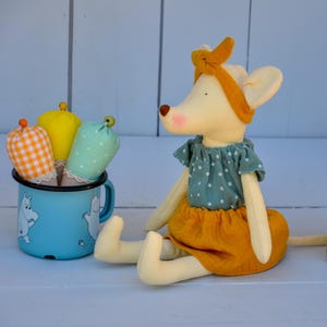 K&ouml;nnte beinhalten: Eine handgefertigte Pl&uuml;schmauspuppe mit gelbem Stirnband, blauem Oberteil mit wei&szlig;en Punkten und senfgelbem Rock. Ein blauer Emaillebecher mit Cartoonfiguren enth&auml;lt drei stoffbezogene Gegenst&auml;nde in orangefarbenem Vichy-Karo, Gelb und blauen Punkten.