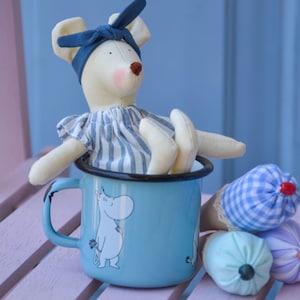 May include: A cream-coloured fabric mouse doll with pink cheeks, wearing a blue headband and a blue and white striped dress, sits inside a light blue enamel mug featuring a white cartoon character illustration. Nearby are small fabric cones.