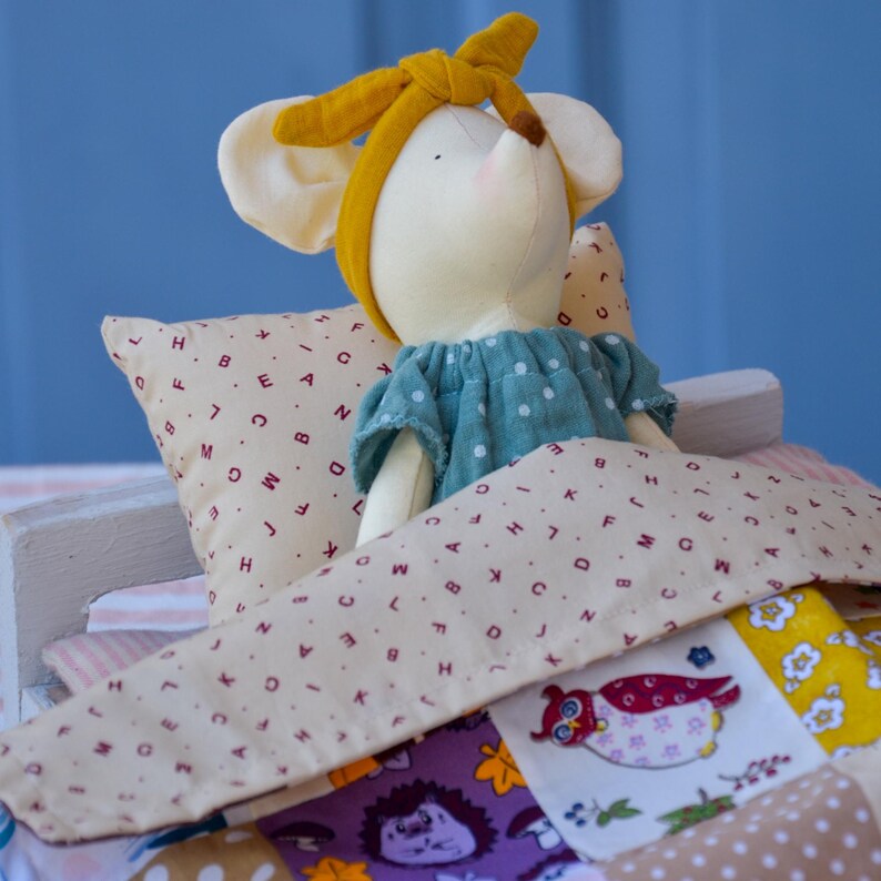K&ouml;nnte beinhalten: Eine Stoffmaus-Puppe mit blauem Kleid und gelbem Stirnband in einem Miniaturbett. Das Bett hat ein Kissen und eine Patchwork-Steppdecke mit verschiedenen Mustern und Farben, darunter eine Eule und ein Igel.