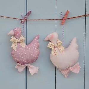 May include: Two fabric goose ornaments, one pink with white polka dots and the other solid pink, are hanging from a clothesline. Each goose has a yellow bow with pink polka dots and a matching bow at the base. The background is a light blue wooden surface.