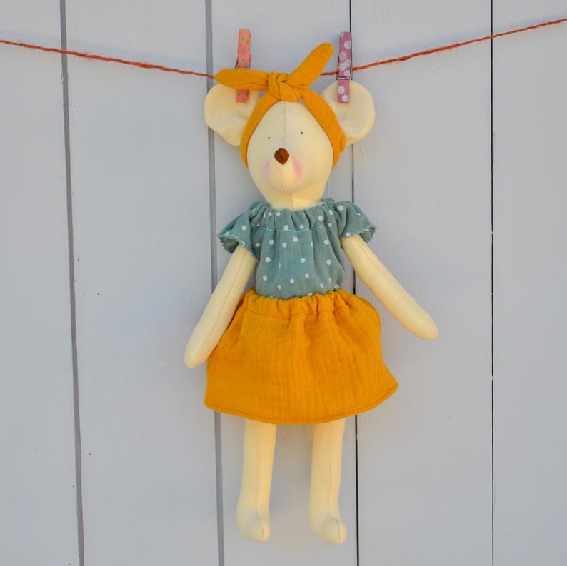 K&ouml;nnte beinhalten: Handgefertigte Stoffpuppe in Form einer Maus, an einer W&auml;scheleine h&auml;ngend. Die Puppe tr&auml;gt ein gelbes Stirnband und ein Kleid mit einem tealfarbenen Oberteil und einem gelben Rock. Sie hat gro&szlig;e, runde Ohren und ein einfaches Gesicht.