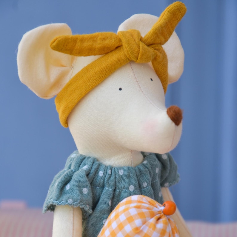 K&ouml;nnte beinhalten: Eine handgefertigte Stoffmaus-Puppe mit wei&szlig;em K&ouml;rper, gro&szlig;en cremefarbenen Ohren und einer braunen Nase. Die Maus tr&auml;gt ein gelbes Stirnband und ein blaues Kleid mit wei&szlig;en Punkten. Sie h&auml;lt eine kleine orange-gelb karierte Tasche.
