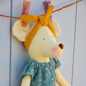 K&ouml;nnte beinhalten: Eine handgefertigte Pl&uuml;schmauspuppe mit gelbem Stirnband und einem tealfarbenen Kleid mit wei&szlig;en Punkten. Die Puppe hat eine rosa Nase und h&auml;ngt an einer W&auml;scheleine mit rosa und lila W&auml;scheklammern.