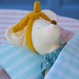 K&ouml;nnte beinhalten: Eine Stoffmaus-Puppe mit gelbem Stirnband, gekleidet in einem blauen gepunkteten Kleid und unter einer blau-wei&szlig; gestreiften Decke. Die Maus hat eine braune Nase und rosige Wangen, was eine sanfte, beruhigende &Auml;sthetik erzeugt.