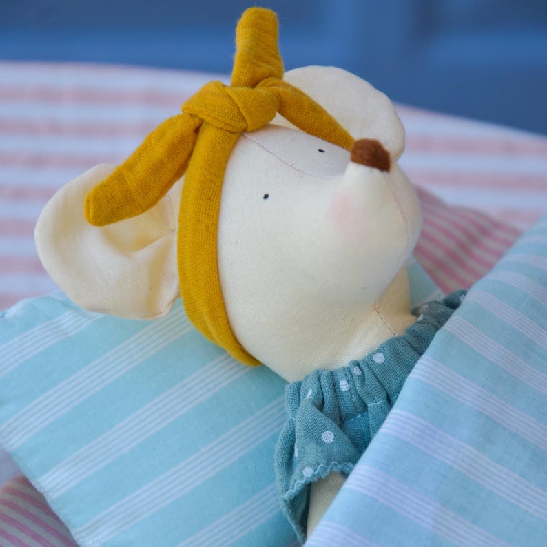K&ouml;nnte beinhalten: Eine Stoffmaus-Puppe mit gelbem Stirnband, gekleidet in einem blauen gepunkteten Kleid und unter einer blau-wei&szlig; gestreiften Decke. Die Maus hat eine braune Nase und rosige Wangen, was eine sanfte, beruhigende &Auml;sthetik erzeugt.