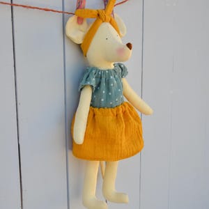 K&ouml;nnte beinhalten: Eine weiche, pl&uuml;schige Mauspuppe mit einem tealfarbenen Oberteil mit Polka-Dots und einem senfgelben Rock. Die Puppe hat ein passendes senfgelbes Stirnband. Die Puppe h&auml;ngt an einer W&auml;scheleine mit rosa W&auml;scheklammern.