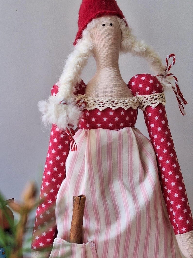 May include: A handmade fabric doll with a red hat and long white hair. The doll's dress features red fabric with white stars and a pink and white striped skirt. A cinnamon stick is in the doll's pocket.