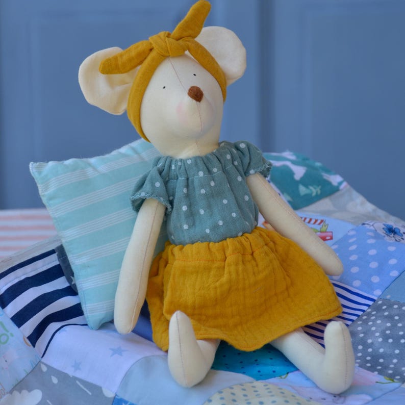 K&ouml;nnte beinhalten: Handgemachte Stoffmaus-Puppe mit gelbem Stirnband, tr&auml;gt ein t&uuml;rkisfarbenes Oberteil mit Punkten und einen gelben Rock. Die Puppe hat cremefarbene Ohren, Arme und Beine und sitzt auf einem Patchwork-Quilt mit blau-wei&szlig;en Mustern.