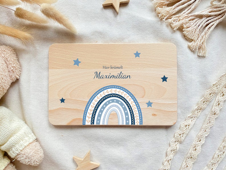 May include: A wooden sign with a blue and white rainbow design and stars. The sign says "Hier kr&uuml;melt Maximilian".