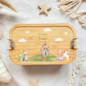 May include: A wooden lunchbox with a light brown finish and silver clasps. The lunchbox has a printed design of a castle, a prince, a princess, a dragon, and a unicorn. The text "Hier krummelt Leonie" is printed on the lunchbox.