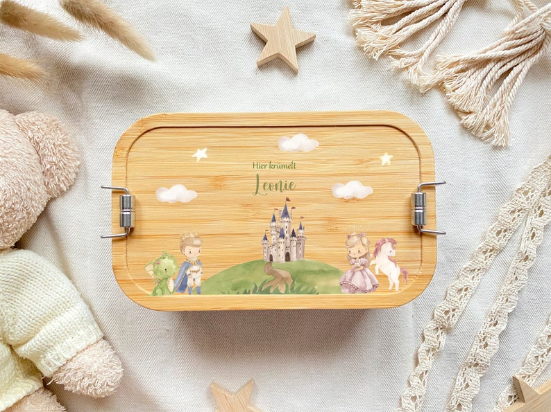 May include: A wooden lunchbox with a light brown finish and silver clasps. The lunchbox has a printed design of a castle, a prince, a princess, a dragon, and a unicorn. The text "Hier krummelt Leonie" is printed on the lunchbox.