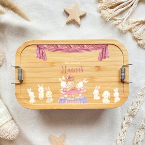 May include: A personalized bamboo lunchbox with a pink and white ballerina and bunnies design. The lunchbox has a silver latch and the text "Her name is Hannah" is printed on the front.