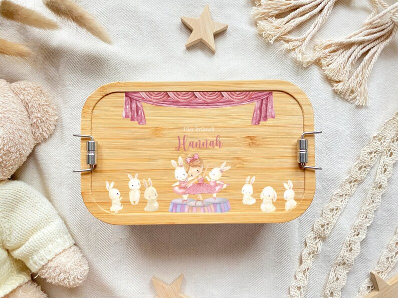 May include: A personalized bamboo lunchbox with a pink and white ballerina and bunnies design. The lunchbox has a silver latch and the text "Her name is Hannah" is printed on the front.