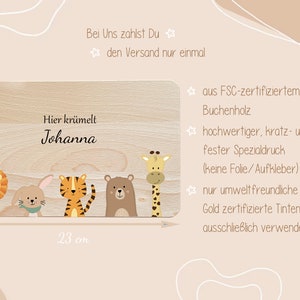 May include: A personalized wooden cutting board with a light wood finish. The board features a cute illustration of a lion, rabbit, tiger, bear, and giraffe. The text "Hier kr&uuml;melt Johanna" is printed on the board. The board measures 23 cm by 15 cm.