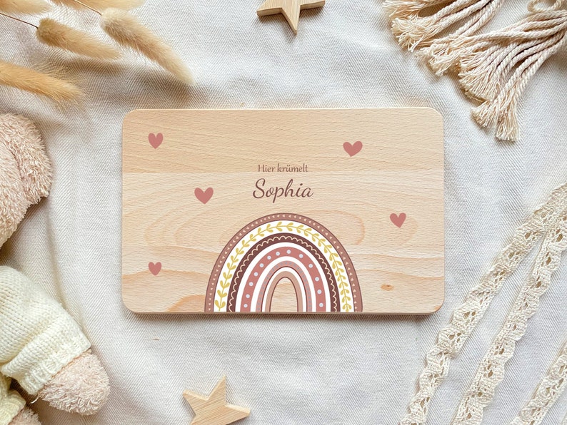 May include: A wooden sign with a rainbow design in shades of pink, brown, and yellow. The sign has the text "Hier kr&uuml;melt Sophia" in German.