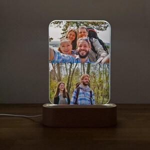 Op de afbeelding: Een houten fotolamp met een helder acryl paneel waarop twee foto's te zien zijn van een groep vrienden in een bosrijke omgeving. De bovenste foto toont vier mensen die lachen en een selfie maken, terwijl de onderste foto twee mensen laat zien die naast elkaar lopen.