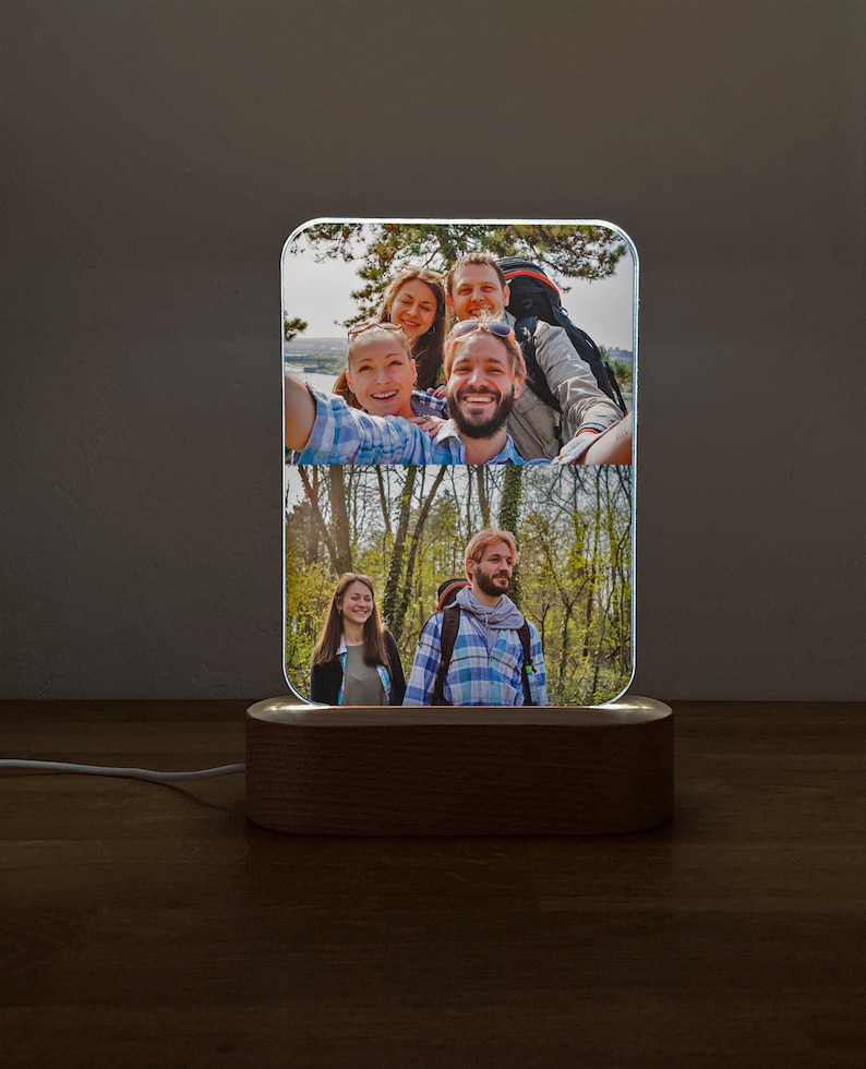 Op de afbeelding: Een houten fotolamp met een helder acryl paneel waarop twee foto's te zien zijn van een groep vrienden in een bosrijke omgeving. De bovenste foto toont vier mensen die lachen en een selfie maken, terwijl de onderste foto twee mensen laat zien die naast elkaar lopen.