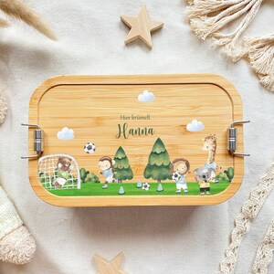 May include: A wooden lunchbox with a light brown finish and a latch closure. The lunchbox has a colorful design featuring animals playing soccer, trees, and clouds. The text "Hier kr&uuml;melt Hanna" is printed on the lunchbox.