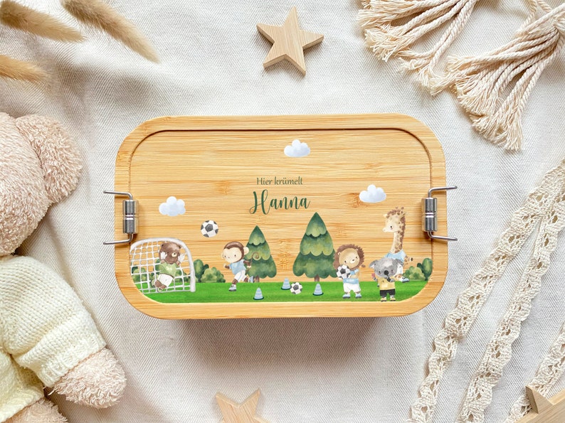 May include: A wooden lunchbox with a light brown finish and a latch closure. The lunchbox has a colorful design featuring animals playing soccer, trees, and clouds. The text "Hier kr&uuml;melt Hanna" is printed on the lunchbox.
