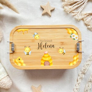 May include: A wooden lunchbox with a bee theme. The lunchbox has a brown wooden exterior with a clear plastic lid. The lunchbox has a beehive, bees, and honeycombs printed on it. The text "Hier kr&uuml;melt Helena" is printed on the lunchbox.