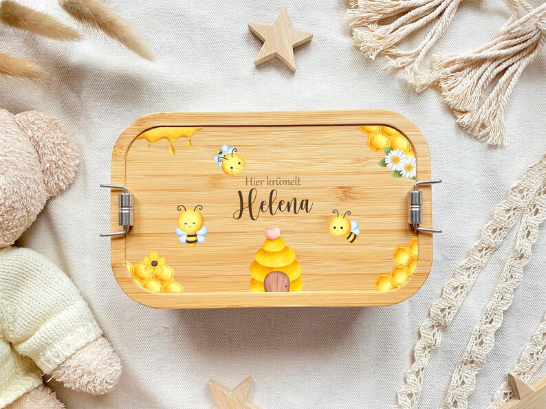 May include: A wooden lunchbox with a bee theme. The lunchbox has a brown wooden exterior with a clear plastic lid. The lunchbox has a beehive, bees, and honeycombs printed on it. The text "Hier kr&uuml;melt Helena" is printed on the lunchbox.