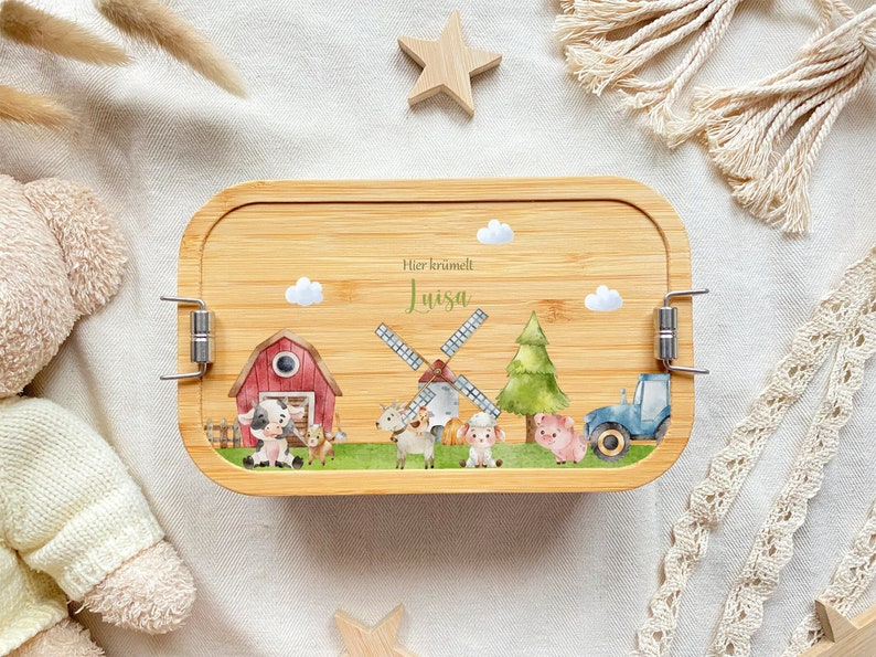 May include: A wooden lunchbox with a farm scene featuring a red barn, windmill, tractor, and animals. The lunchbox has a personalized inscription that reads "Hier kr&uuml;melt Luisa".