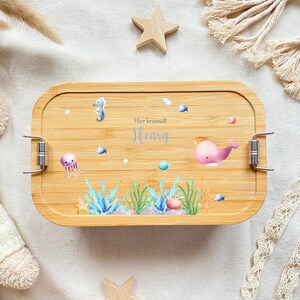 May include: A light brown bamboo lunch box with a blue and pink sea creature design. The lunch box has a silver latch and the text "Hier kr&uuml;melt Henry" is printed on the front.