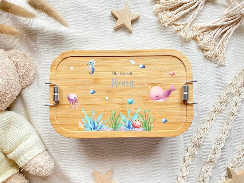May include: A light brown bamboo lunch box with a blue and pink sea creature design. The lunch box has a silver latch and the text "Hier kr&uuml;melt Henry" is printed on the front.