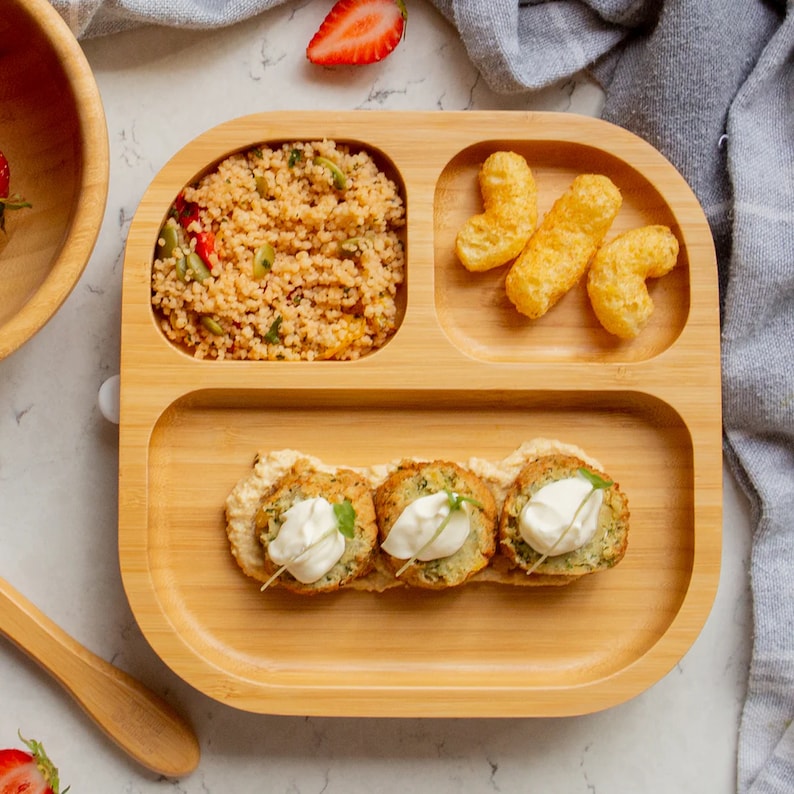 Peut inclure: Une assiette divis&eacute;e en bois avec quatre sections. La section sup&eacute;rieure gauche contient une portion de couscous avec des l&eacute;gumes. La section sup&eacute;rieure droite contient quelques en-cas de fromage souffl&eacute;. La section inf&eacute;rieure contient trois aliments pan&eacute;s et frits avec une cuiller&eacute;e de sauce blanche sur le dessus.