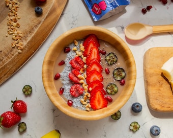 May include: A wooden bowl filled with a chia pudding topped with sliced strawberries, raspberries, blueberries, pomegranate seeds, and chopped hazelnuts.
