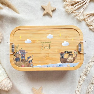 May include: A wooden lunchbox with a light brown finish and a latch closure. The lunchbox features a whimsical illustration of pirates on a boat with a parrot, stars, and clouds. The text "Hier kr&uuml;melt Emil" is printed on the lunchbox.