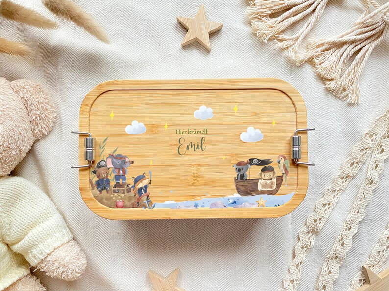 May include: A wooden lunchbox with a light brown finish and a latch closure. The lunchbox features a whimsical illustration of pirates on a boat with a parrot, stars, and clouds. The text "Hier kr&uuml;melt Emil" is printed on the lunchbox.