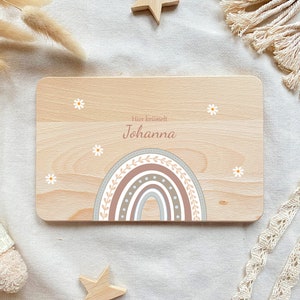 May include: A wooden sign with a rainbow design and the text "Hier  braumelt Johanna" in pink lettering. The rainbow is decorated with brown leaves and white flowers.