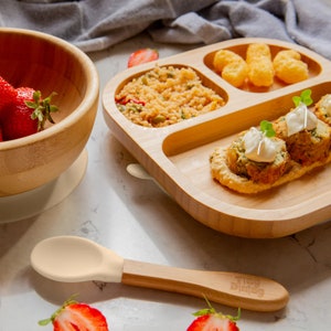 Peut inclure: Une assiette divis&eacute;e en bois avec quatre sections, remplie de nourriture. Les sections contiennent une salade verte, une sauce blanche avec des rouleaux de viande, une sauce jaune avec des rouleaux de viande et un tas de p&eacute;pites dor&eacute;es. Une cuill&egrave;re en bois avec une pointe en silicone jaune clair est au premier plan. L'assiette a une bordure en silicone blanc.