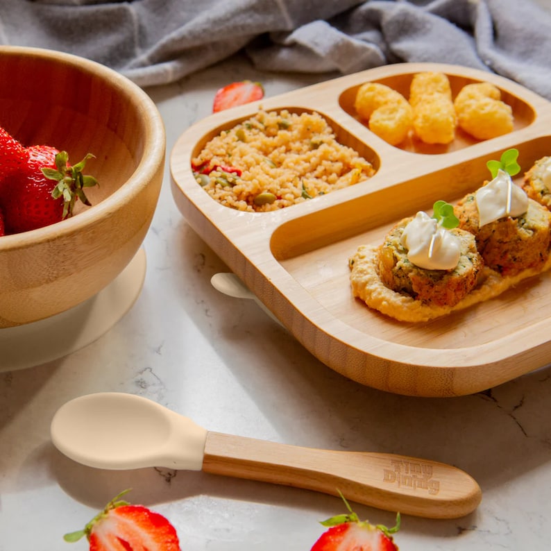 Peut inclure: Une assiette divis&eacute;e en bois avec quatre sections, remplie de nourriture. Les sections contiennent une salade verte, une sauce blanche avec des rouleaux de viande, une sauce jaune avec des rouleaux de viande et un tas de p&eacute;pites dor&eacute;es. Une cuill&egrave;re en bois avec une pointe en silicone jaune clair est au premier plan. L'assiette a une bordure en silicone blanc.