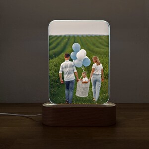 Op de afbeelding: Een houten basis met een helder acryl paneel waarop een foto te zien is van een gezin van drie dat in een veld loopt. De foto toont een man, een vrouw en een kind dat ballonnen vasthoudt. Het acryl paneel wordt van achteren verlicht.