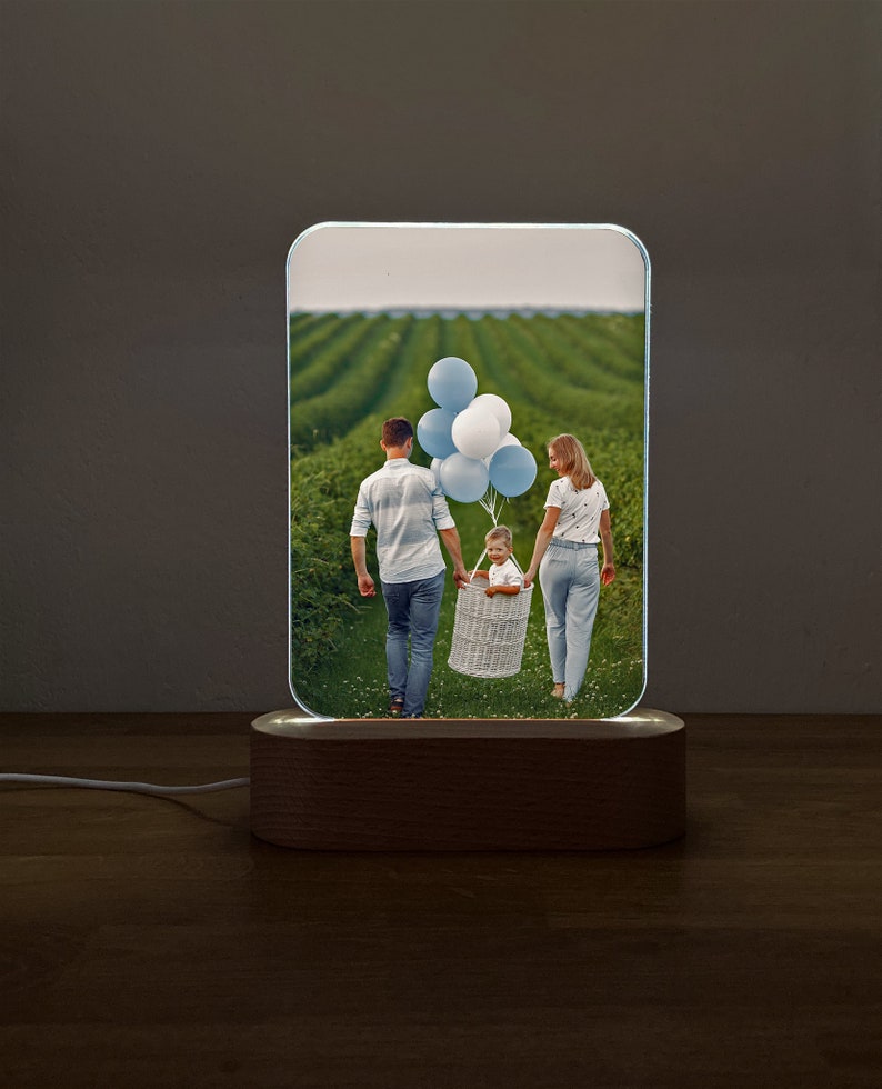 Op de afbeelding: Een houten basis met een helder acryl paneel waarop een foto te zien is van een gezin van drie dat in een veld loopt. De foto toont een man, een vrouw en een kind dat ballonnen vasthoudt. Het acryl paneel wordt van achteren verlicht.