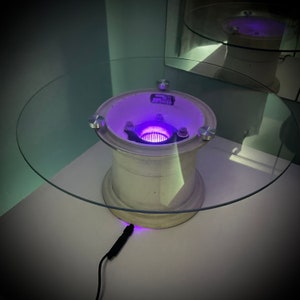 Peut inclure: Une table basse en verre avec un piétement unique fabriqué à partir d'une jante de pneu de voiture réutilisée. La jante a une lumière violette qui brille à travers le trou central. La table est sur une surface blanche et a un cordon noir branché.