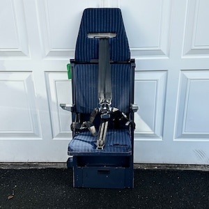 May include: A blue aeroplane seat with a black safety belt and a small storage compartment underneath. The seat is made of a striped fabric and has a padded back and seat.