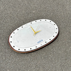 May include: A silver oval clock with a gold hand and black numbers. The clock face has the text "656CB" printed on it.